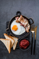 Fried eggs on a stylish ceramic plate with handles with fried bacon, cherry tomatoes, tomato paste and bread toast on a dark background. Breakfast, lunch and dinner. Delicious food.