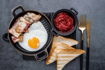 Fried eggs on a stylish ceramic plate with handles with fried bacon, cherry tomatoes, tomato paste and bread toast on a dark background. Breakfast, lunch and dinner. Delicious food.