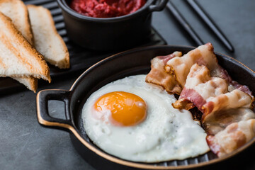 Fried eggs on a stylish ceramic plate with handles with fried bacon, cherry tomatoes, tomato paste and bread toast on a dark background. Breakfast, lunch and dinner. Delicious food.