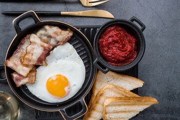 Fried eggs on a stylish ceramic plate with handles with fried bacon, cherry tomatoes, tomato paste and bread toast on a dark background. Breakfast, lunch and dinner. Delicious food.