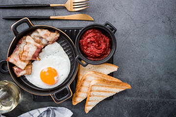 Fried eggs on a stylish ceramic plate with handles with fried bacon, cherry tomatoes, tomato paste and bread toast on a dark background. Breakfast, lunch and dinner. Delicious food.