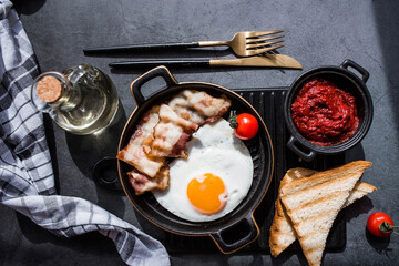 Fried eggs on a stylish ceramic plate with handles with fried bacon, cherry tomatoes, tomato paste and bread toast on a dark background. Breakfast, lunch and dinner. Delicious food.