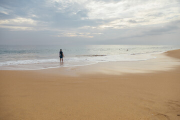 Young boy with a surfboard standing on the sandy ocean beach in the sunset time.