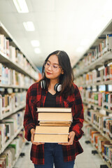 Smart Asian female student in casual style bring pile of books in library while looking for another books at the bookshelf. 