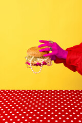 Portrait of female hands of rich woman wearing gold rings holding big burger over yellow background. Trash pop art, bright colors