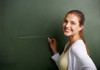 Ready to enrich young minds. A beautiful young teacher writing on a blackboard while smiling at the camera.