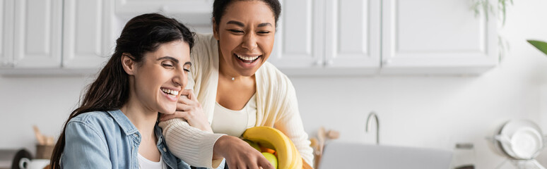 joyful multiracial woman holding bowl with fruits near girlfriend at home, banner.