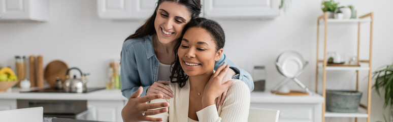 happy interracial lesbian couple hugging each other in kitchen, banner.