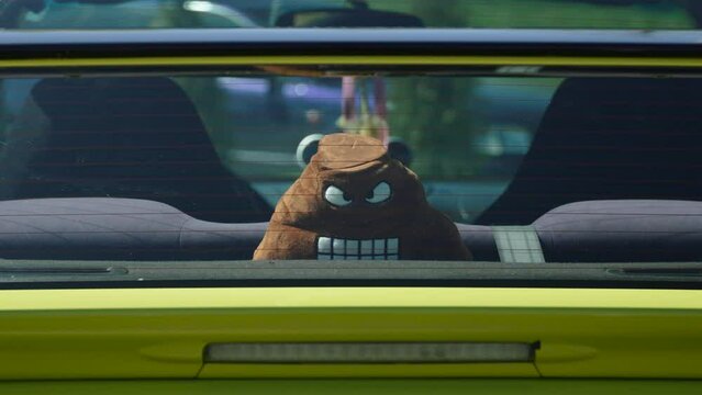Plush toy in form of an evil pile of poop lies in cabin near rear window of yellow sports car, close-up. Sunny summer day in car park with trees.