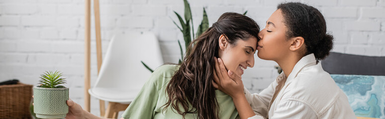 joyful multiracial woman kissing forehead of happy girlfriend with green plant, banner.