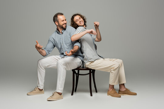 smiling father and teenage son having fun and pushing each other while sitting on same chair on grey.