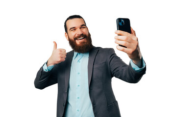 Handsome bearded man is taking a selfie with his phone while showing thumb up over white background.
