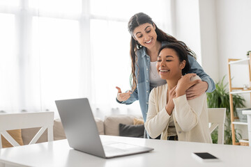 happy lesbian woman pointing at multiracial girlfriend near laptop during video call.