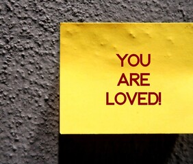 Yellow note on wall with handwritten text - YOU ARE LOVED - supportive phrase to encourage someone...