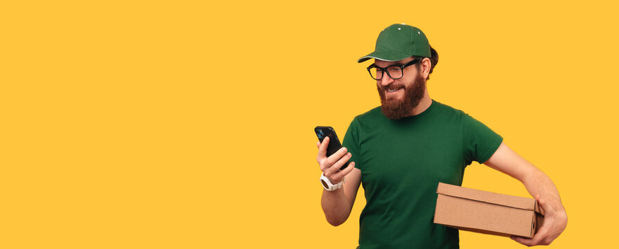 Banner shot of a man holds a box he has to deliver while looking to his phone over yellow background.