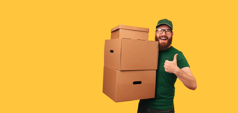 Banner shot of an excited delivery man holding some boxes and a thumb up over yellow background.