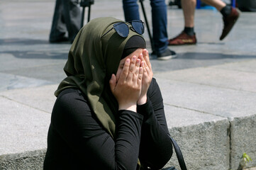 Young Muslim woman in hijab and sunglasses feeling shy covering face with both hands sitting on the pavement on street