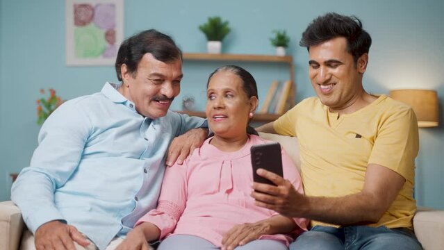 Happy Indian adult son showing mobile phone to senior parents while sitting on sofa at home - concept of matrimony app, technology and family bonding