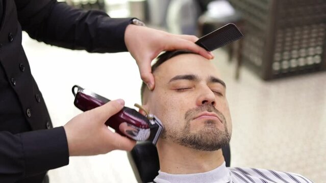 stylish barber cutting beard of client with trimmer while working in barbershop
