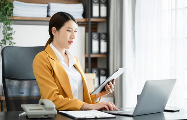 young asian businesswoman using tablet surfing the internet, searching information during online working on laptop computer at home office.