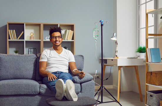 Young happy african american man sitting on the couch in the clinic while receiving IV drip infusion and vitamin therapy in his blood. Cheerful smiling person receiving injection therapy.