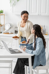 multiracial woman holding plates with baked croissants near happy girlfriend working on laptop from home.