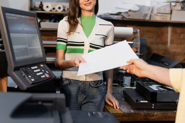 cropped view of happy publisher giving blank paper to colleague in print center.