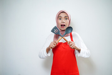 Cheerful asian muslim woman in red apron holding soup spoon and spatula. Cooking ware. Cooking concept.