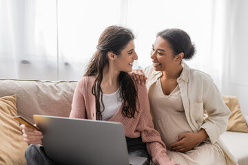 happy lesbian woman holding credit card while doing online shopping with cheerful and pregnant multiracial partner.