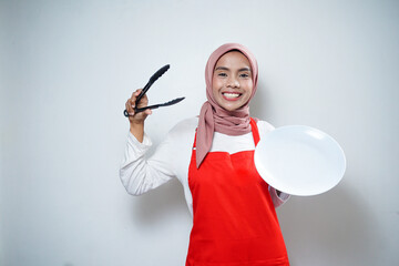 Cheerful asian muslim woman in red apron holding empty plate and tongs. Food Advertisement. Cooking concept.