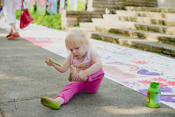 A cheerful cute girl draws with green paints on her leg, the foot of her body. Toddler girl plays...