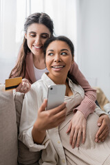 happy and pregnant multiracial woman holding smartphone while doing online shopping with lesbian partner.