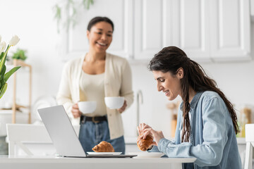 happy lesbian woman eating croissant near laptop and multiracial girlfriend with cups on blurred background.