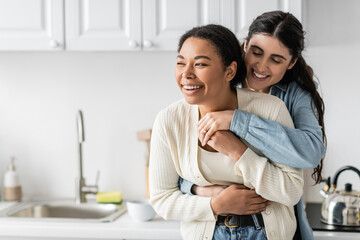 cheerful lesbian woman hugging joyful multiracial girlfriend in modern kitchen.