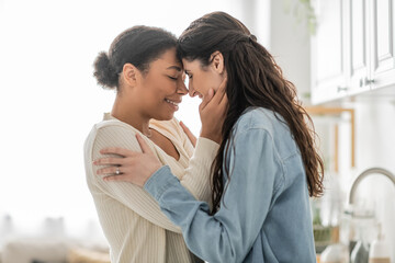 curly multiracial woman hugging cheerful girlfriend with closed eyes at home.