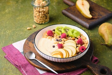 Breakfast, granola with yogurt, pear, kiwi and raspberries in a flat white plate on a green concrete background. Healthy quick breakfasts.