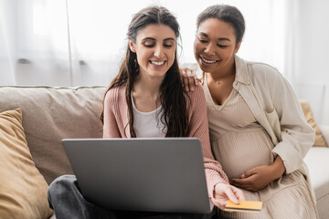 cheerful and pregnant multiracial woman looking at laptop while doing online shopping with happy lesbian partner.