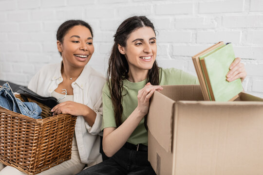 joyful lesbian woman unpacking books from carton box while sitting on sofa next to multiracial girlfriend.