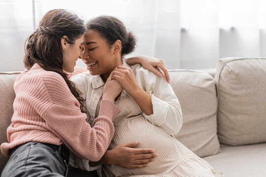 positive lesbian woman hugging pregnant multiracial partner and sitting on couch.