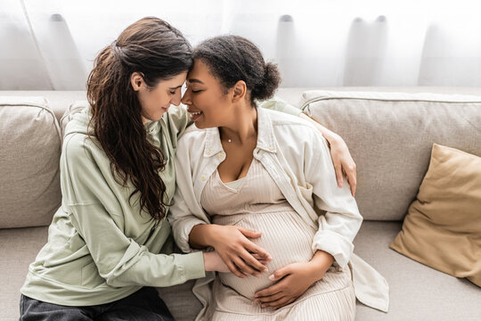 happy lesbian woman hugging pregnant multiracial wife while sitting on sofa.