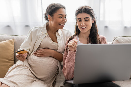 pregnant multiracial woman holding credit card while doing online shopping with cheerful lesbian partner. - Powered by Adobe