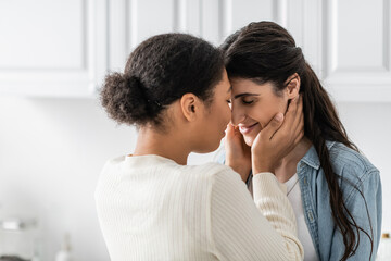 curly multiracial woman hugging happy girlfriend with closed eyes at home.