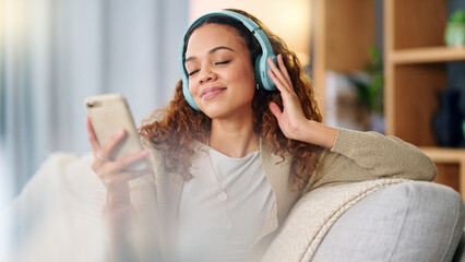 Woman wearing headphones and using a phone to listen to her music while relaxing on a sofa at home. Carefree female dancing and having fun alone on a couch, enjoying her free time on a weekend