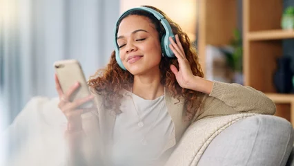 Foto op Plexiglas Woman wearing headphones and using a phone to listen to her music while relaxing on a sofa at home. Carefree female dancing and having fun alone on a couch, enjoying her free time on a weekend © N Felix/peopleimages.com