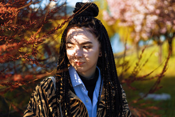 Young tender brunette girl with dreadlocks near herbal branch in front of her face. Portrait of a woman with artistic shadow drops on her face in spring garden.