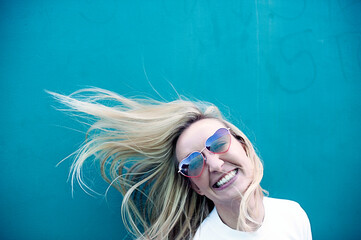 Photo portrait of a young happy woman wearing heart sunglasses next to a blue wall. 