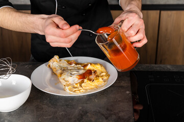 A Cook in a black uniform in the kitchen with ingredients prepared for cooking. Chef making an omelet with bacon for breakfast.