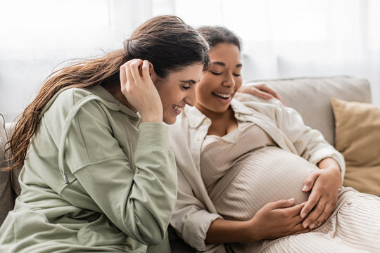 happy lesbian woman smiling while hugging pregnant multiracial wife while sitting on sofa.