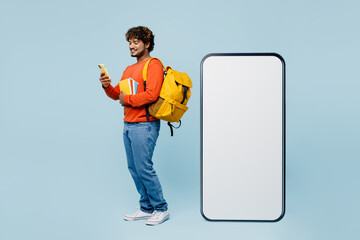 Full body young boy student wear casual clothes backpack bag hold book big huge blank screen mobile cell phone use smartphone isolated on plain blue background High school university college concept.