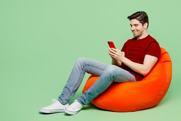 Full body smiling young man he wears red t-shirt casual clothes sit in bag chair hold in hand use...